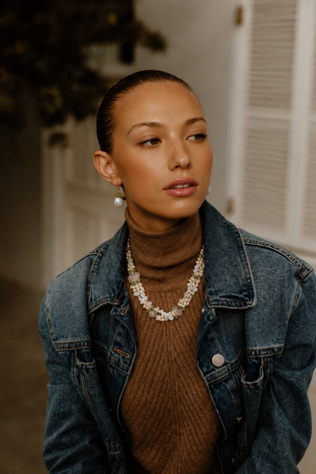 Woman in oversized washed denim jacket over a tan turtleneck, wearing a multi-strand mixed pearl necklace and a single pearl drop earring