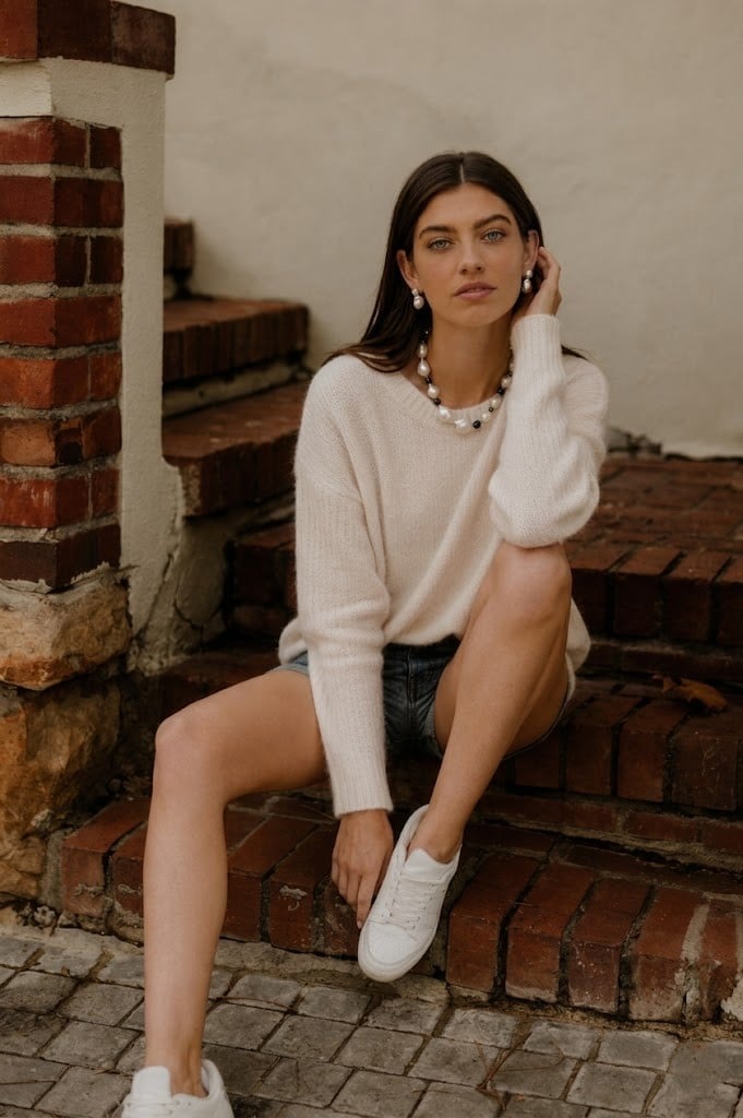 Woman in cream oversized knit, denim shorts and white sneakers seated on red brick steps, wearing a single-strand pearl necklace and pearl drop earrings
